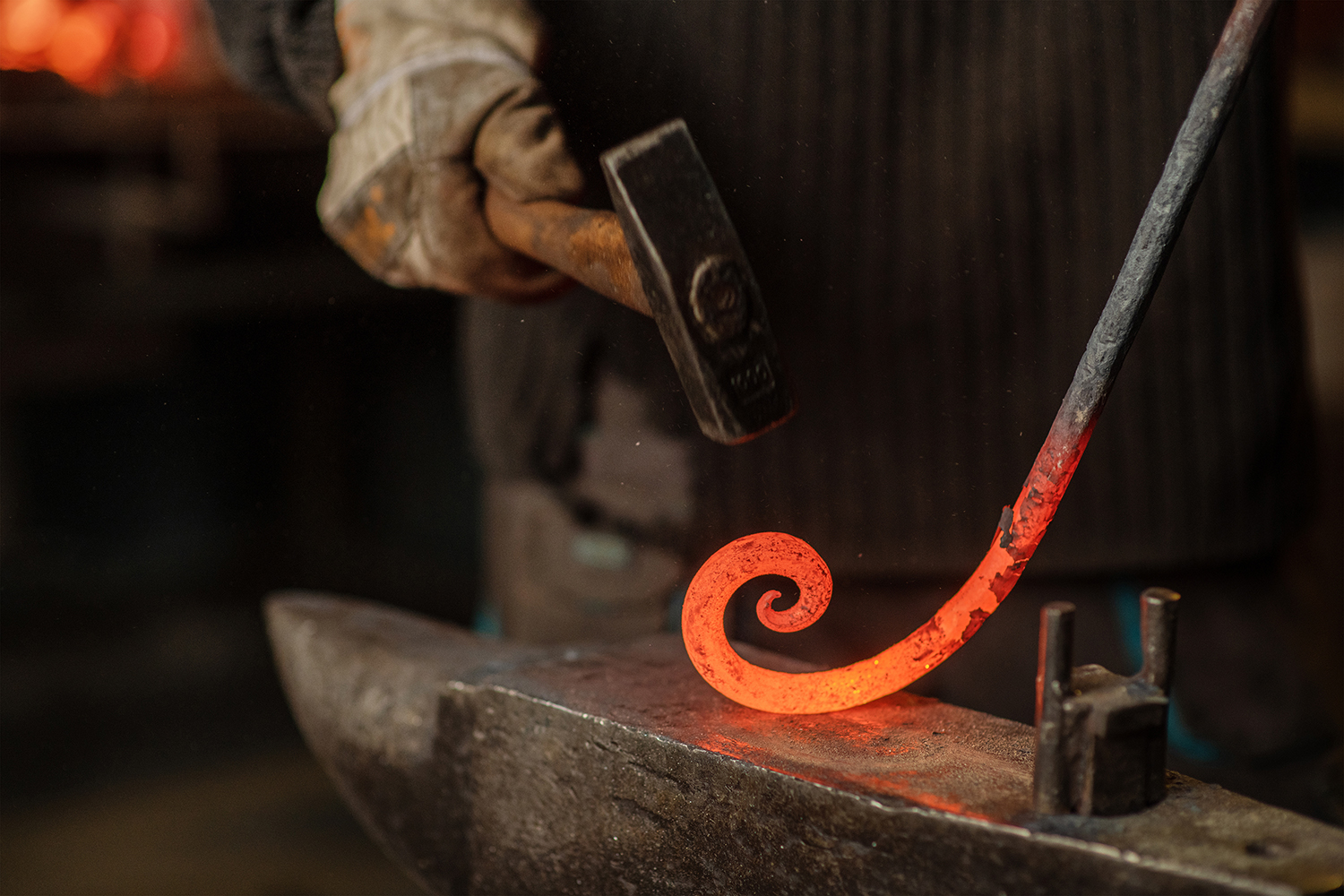 The blacksmith hits the red-hot workpiece in the forge with a hammer and glowing sparks fly in all directions